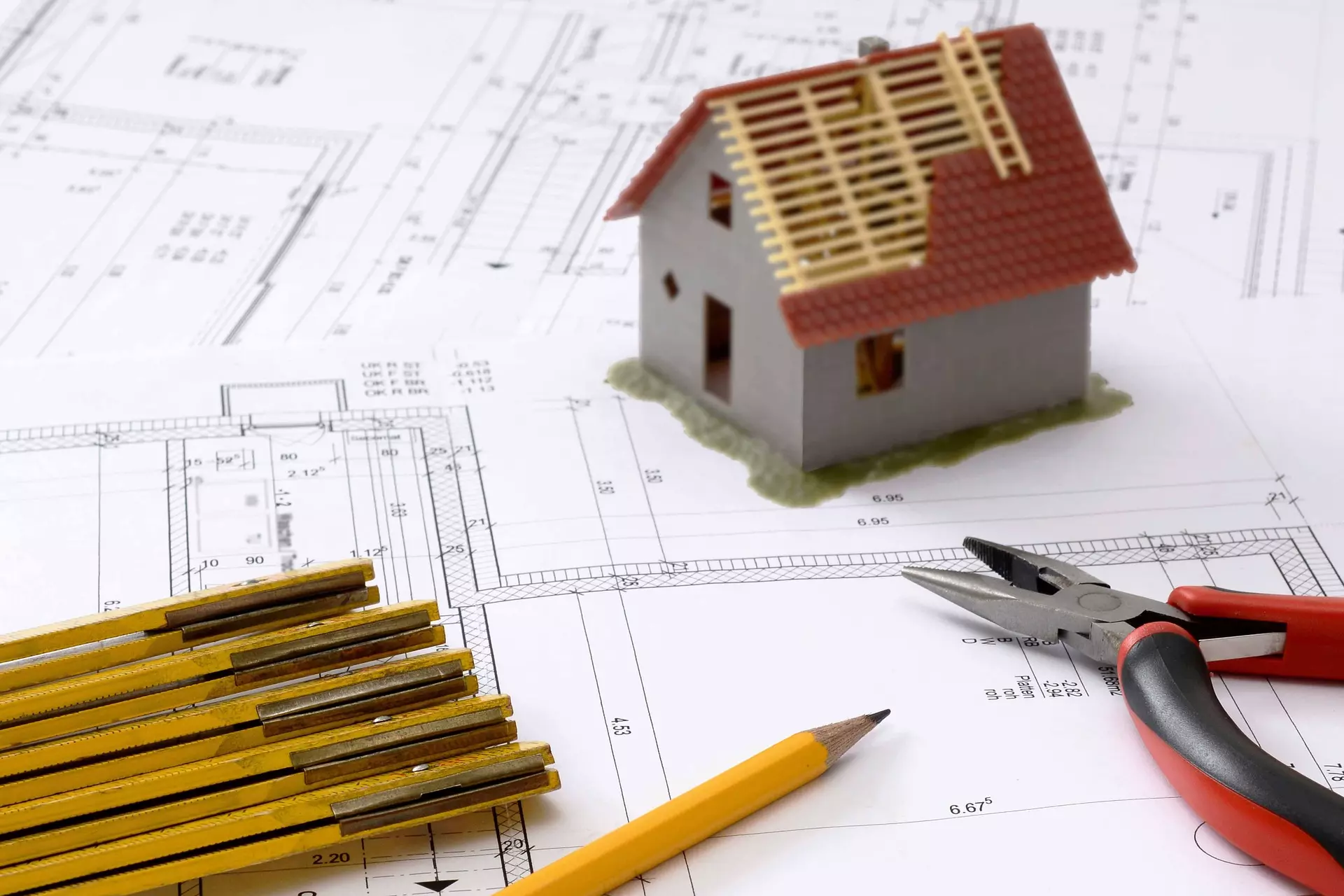 Bild zeigt im Hintergrund einen Plan, darauf ein Haus, eine Zange, Zollstab und einen Bleistift
